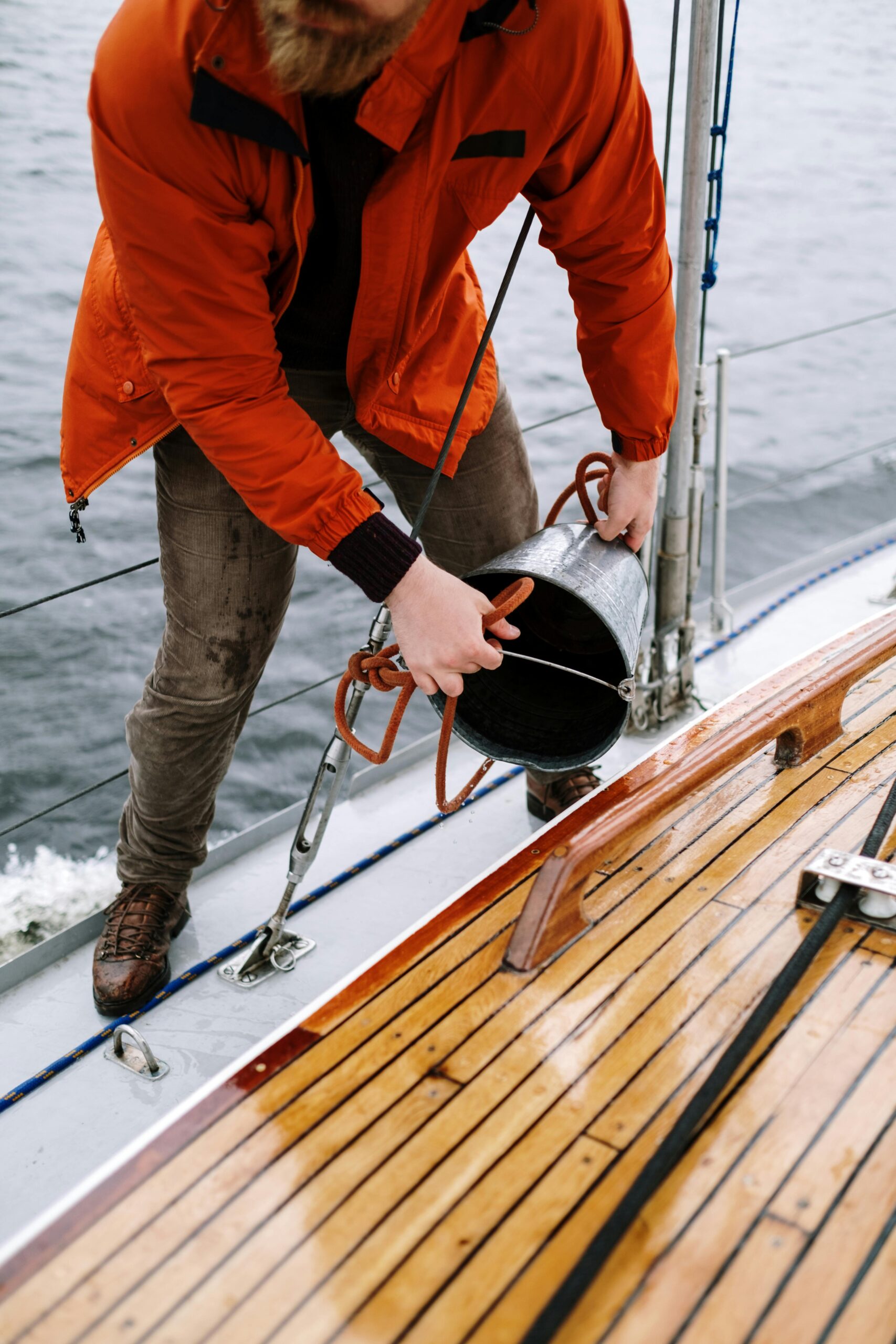 Man in orange jacket cleaning sailboat deck with a bucket on the ocean.